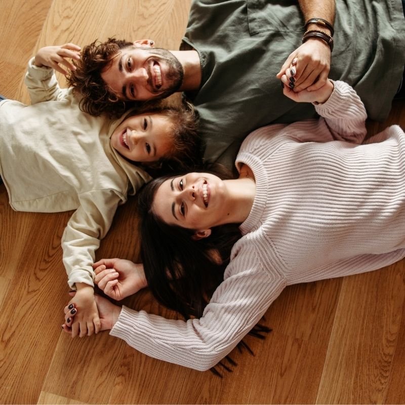 A family lies together on the floor, smiling and enjoying each other's company in a cozy living room setting.