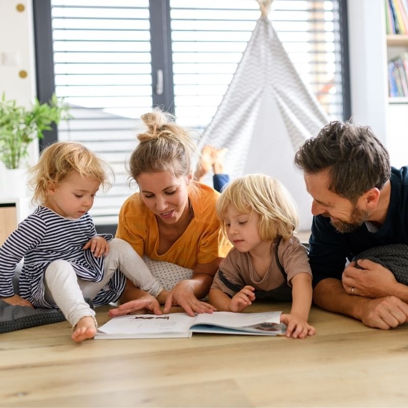 A family of four sits together on a couch, reading a book and sharing smiles, creating a warm, cozy atmosphere.