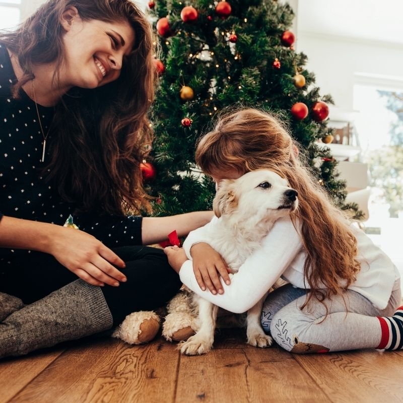 A woman and child sit on the floor, surrounded by gifts, near a decorated Christmas tree.